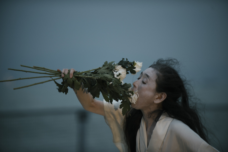 A side view of Eiko who holds up a bouquet of white flowers to her nose